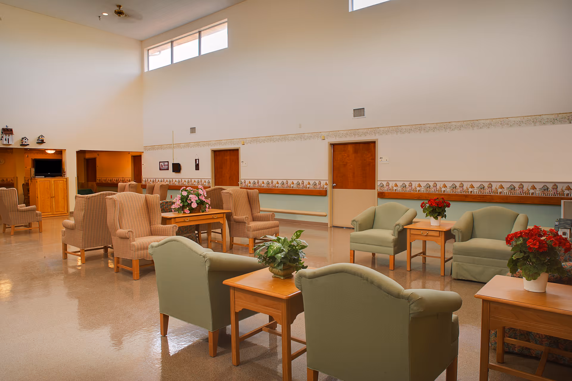 A spacious senior living facility common area with multiple armchairs and small wooden tables arranged in groups. The room has high ceilings with windows near the top, light-colored walls with a decorative border, and several potted plants and flowers on the tables. There is a wooden cabinet with a TV in the background and several closed doors along the wall.