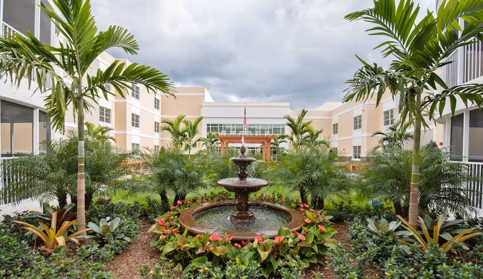 Three-tier fountain in a landscaped courtyard with palm trees and a surrounding multi-story building under a cloudy sky.