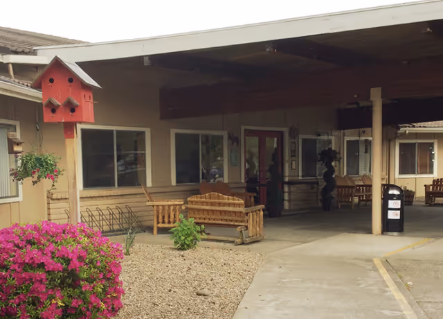 Covered entrance of a nursing facility with wooden benches, a red birdhouse, flowering shrubs, and a driveway.