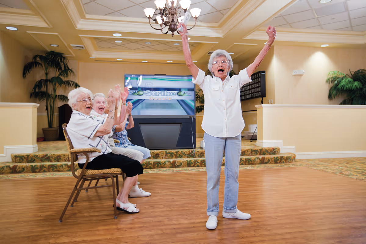 Three elderly women in a spacious, well-lit room with beige walls and carpeted steps. Two women are seated on chairs clapping and smiling, while a third woman stands in front with her arms raised in celebration. A large screen in the background displays a bowling game called 'Strike'.