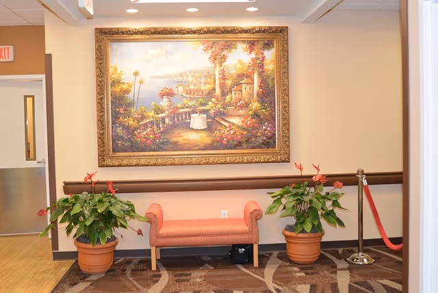 A hallway area in a senior living facility with a coral-colored cushioned bench between two large potted plants with green leaves and red flowers. Above the bench hangs a large framed painting depicting a scenic garden terrace overlooking a coastal village. The walls are beige, and the floor has a patterned carpet. There is an exit sign and a door visible to the left, and a red rope barrier to the right.