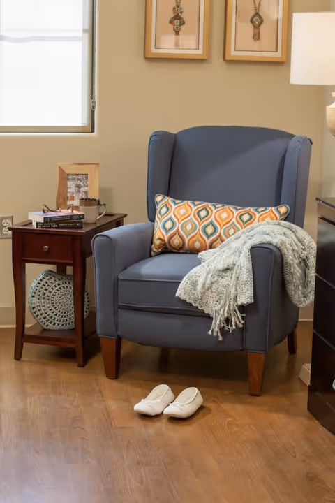 A cozy corner featuring a blue upholstered armchair with a colorful patterned pillow and a light green knitted throw draped over one arm. In front of the chair on the wooden floor are a pair of white slippers. To the left of the chair is a wooden side table with a drawer, holding a framed photo, a mug, and some books. A decorative woven basket is placed on the lower shelf of the table. The wall behind has two framed artworks and a window with a white shade.