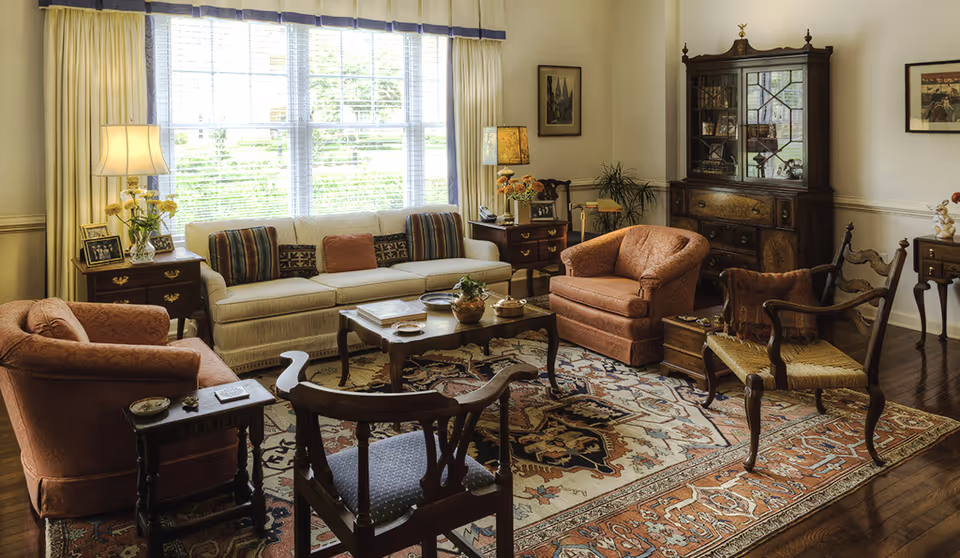 Sunlit traditional living room with a sofa, upholstered armchairs, wooden chairs, a coffee table, patterned rug, and a china cabinet.