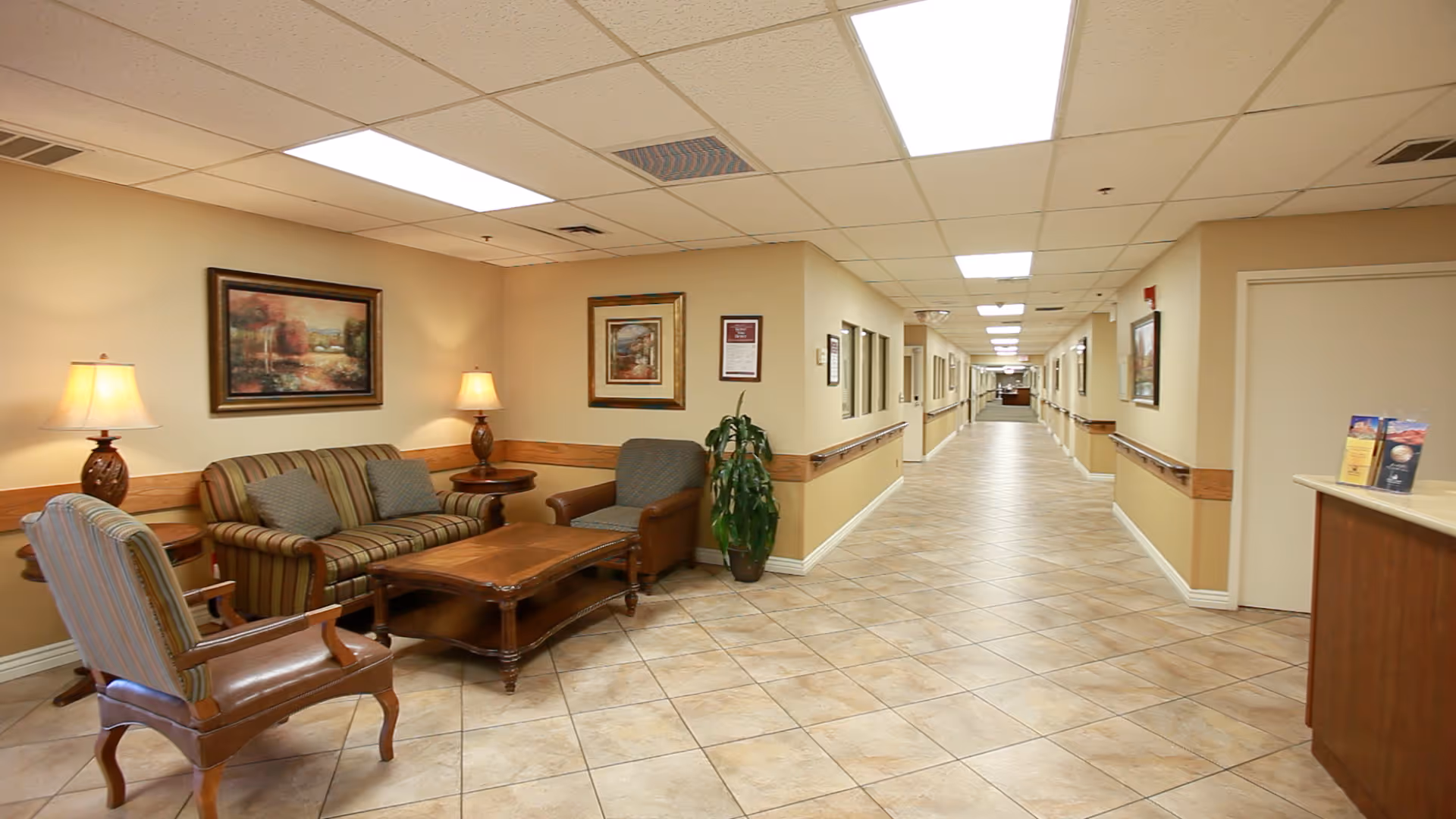 A well-lit hallway in a nursing center with beige walls and tiled floors. On the left side, there is a sitting area with a striped sofa, two armchairs, a wooden coffee table, two table lamps, and framed artwork on the walls. The hallway extends into the distance with doors and handrails along both sides.