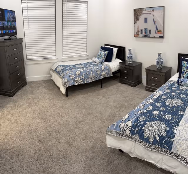 Carpeted bedroom with two twin beds in blue floral bedding, matching nightstands and dresser with a TV beneath two windows.