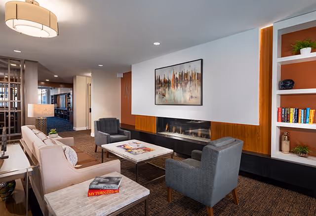 A cozy senior living facility lounge area with a beige sofa, two gray armchairs, a marble coffee table, and a fireplace with a modern abstract painting above it. There are built-in shelves with books and decorative items on the right side, and a lamp on a side table next to the sofa. The room has warm lighting and a carpeted floor.