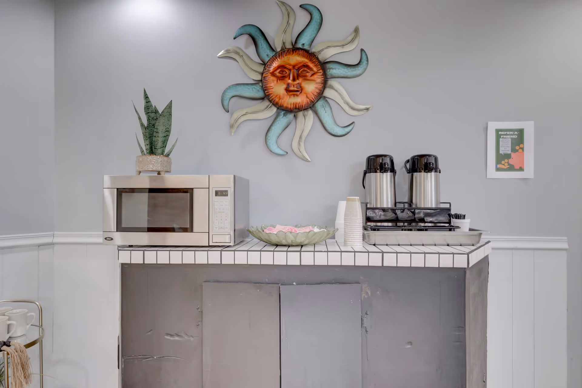 A small refreshment area with a microwave, a potted plant, two coffee dispensers, disposable cups, and a bowl with pink napkins on a tiled countertop. Above the countertop is a decorative sun wall art and a small framed sign on the wall.