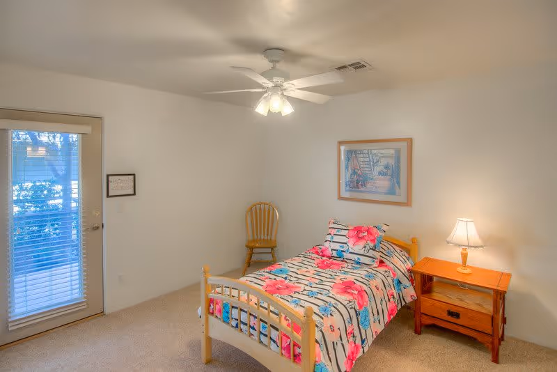 A small bedroom with a single wooden bed covered with a floral bedspread in pink, blue, and white. Next to the bed is a wooden nightstand with a lamp on it. A wooden chair is placed in the corner near a door with glass panels and blinds. A ceiling fan with lights is mounted on the ceiling, and a framed picture hangs on the wall above the bed.