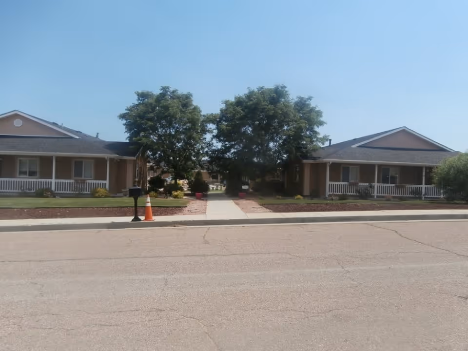 Front view of two single-story assisted living houses with porches, a central sidewalk lined by trees, and a mailbox with an orange traffic cone.