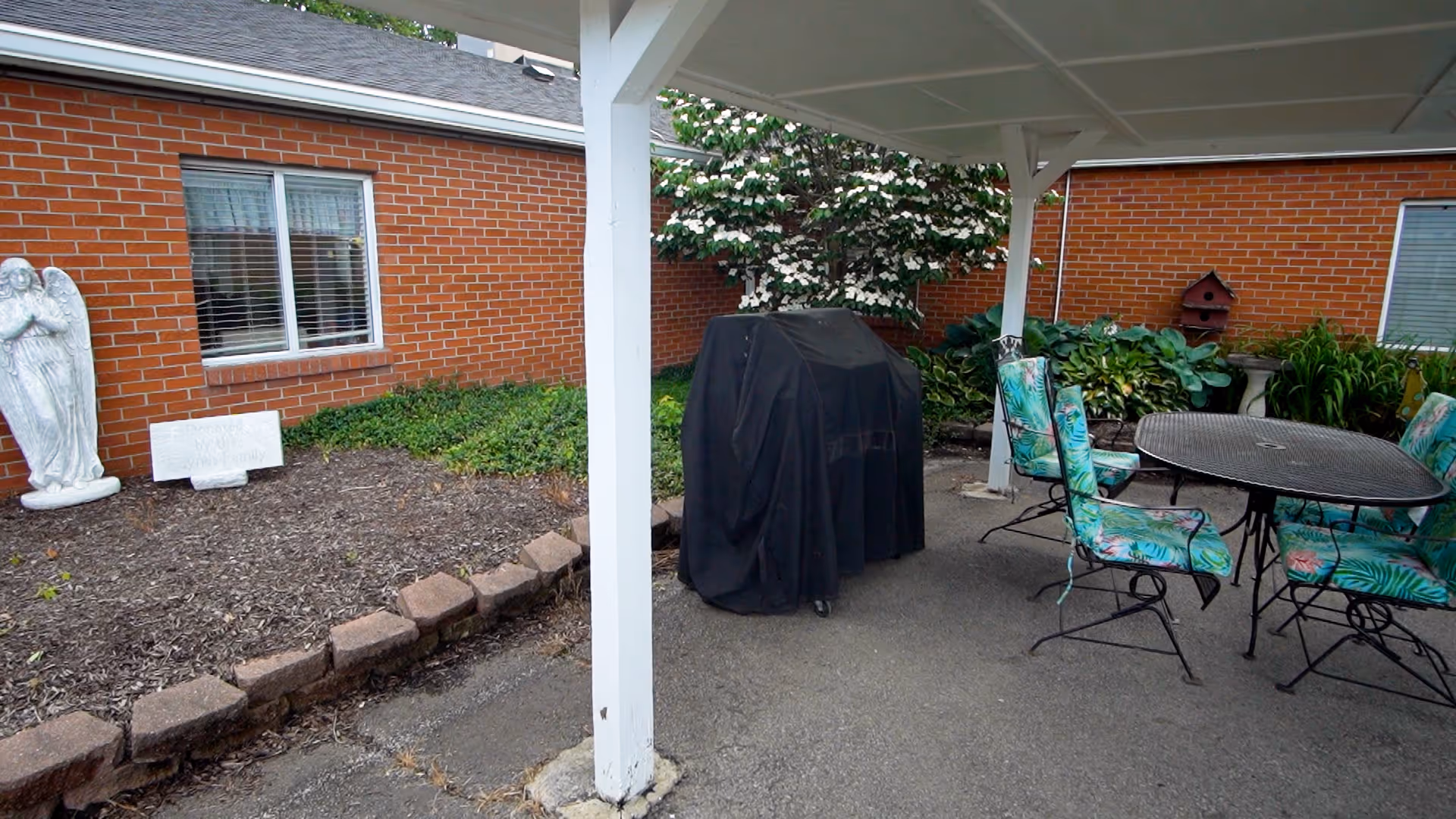 Outdoor patio area with a covered seating space featuring a round metal table and four chairs with floral cushions. A covered grill is positioned near the center. Surrounding the patio are brick walls with windows, garden beds with plants, a white angel statue, and a birdhouse mounted on the wall.