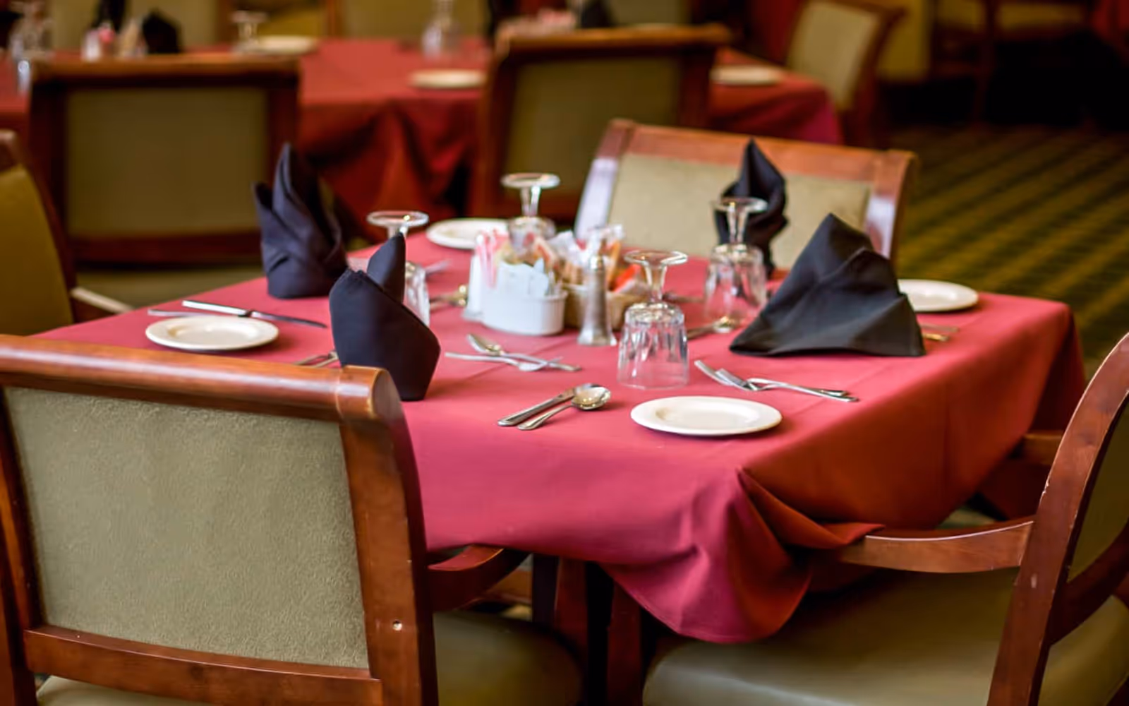 A dining table set with a red tablecloth, black folded napkins, upside-down wine glasses, white plates, and silverware in a senior living facility dining room.