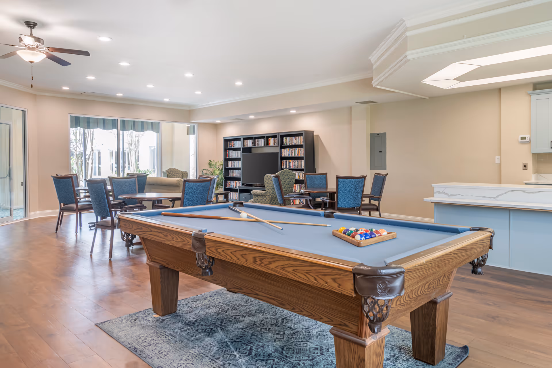 A bright communal game room featuring a pool table in the foreground, tables and chairs, and a TV with shelving in the background.