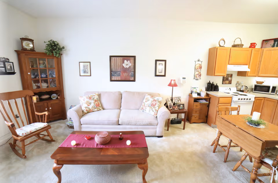 A cozy living room and kitchen area with a beige sofa adorned with floral pillows, a wooden rocking chair with a floral cushion, a wooden coffee table with decorative items on a red runner, and a wooden dining table with chairs. The kitchen features wooden cabinets, a white stove, microwave, and various kitchen items. The walls are decorated with framed pictures and a small lamp on a side table next to the sofa.