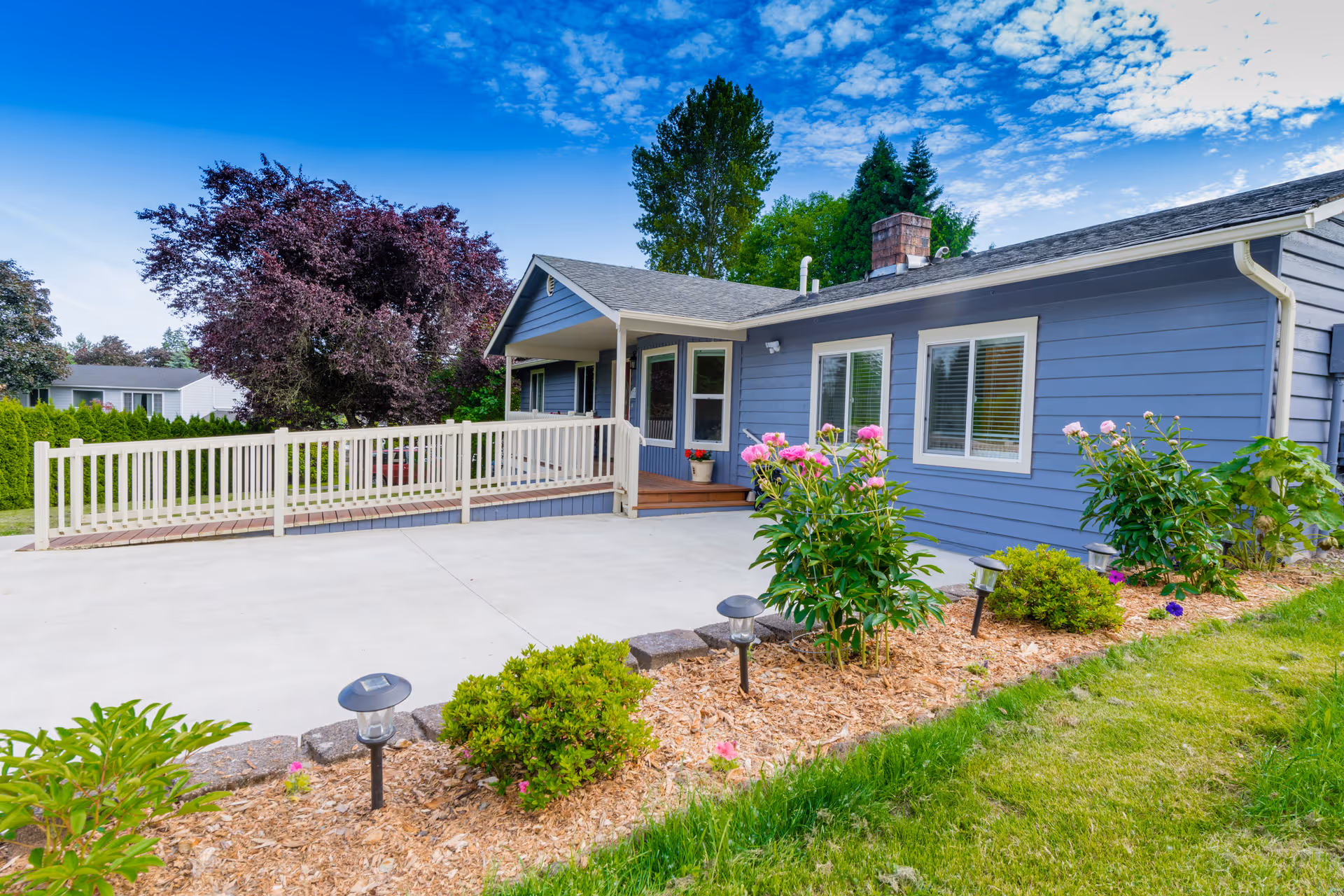 Exterior view of a single-story blue house with white trim, a concrete driveway, a white railing ramp leading to the front door, and a garden bed with green shrubs and pink flowers under a partly cloudy blue sky.