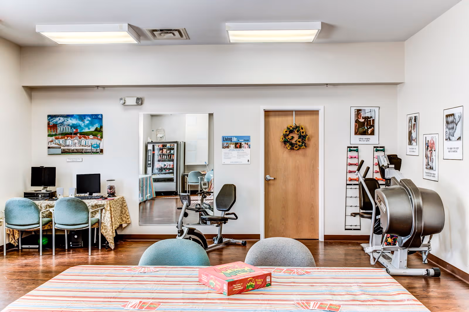 A multi-purpose room in a senior living facility with a table covered by a striped tablecloth and a board game on top. In the background, there are exercise machines, a computer desk with two computers, chairs, a door decorated with a wreath, and various framed pictures and posters on the walls.