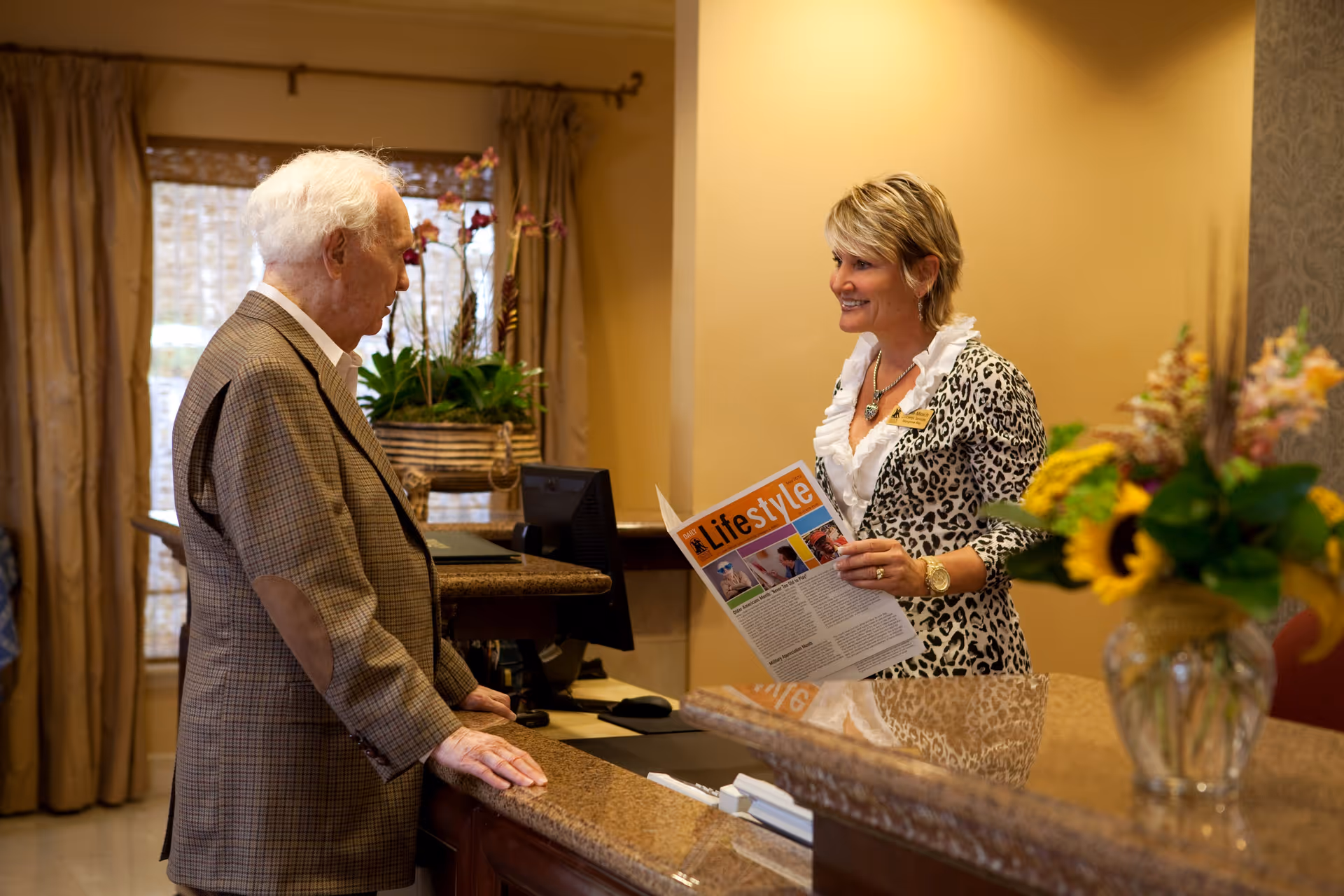 An elderly man in a brown plaid jacket is standing at a reception desk talking to a smiling woman behind the counter who is holding a magazine titled 'Lifestyle'. The reception area has a granite countertop, a vase with flowers, and a window with curtains in the background.