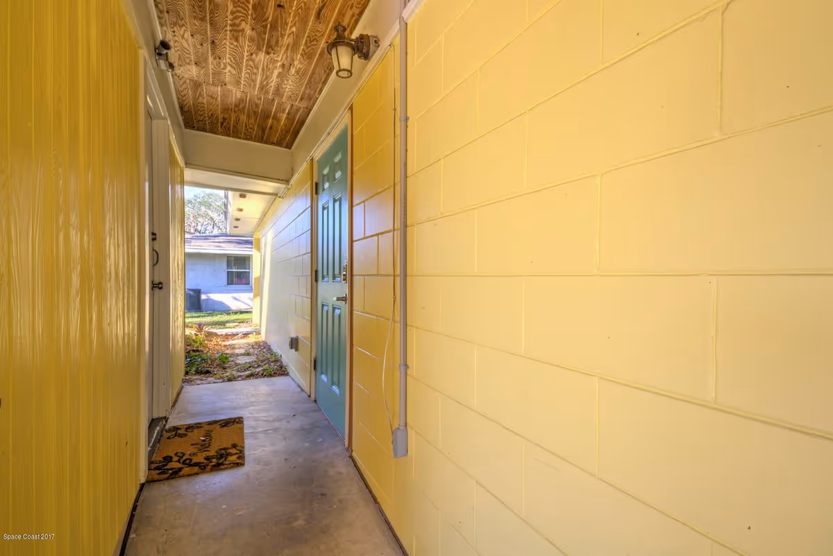 Narrow yellow exterior hallway with green door, wood ceiling, and concrete floor.