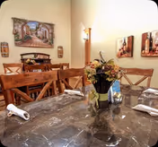 Marble-top dining table with a floral centerpiece, folded napkins and wooden chairs in a warmly lit dining room with wall art.