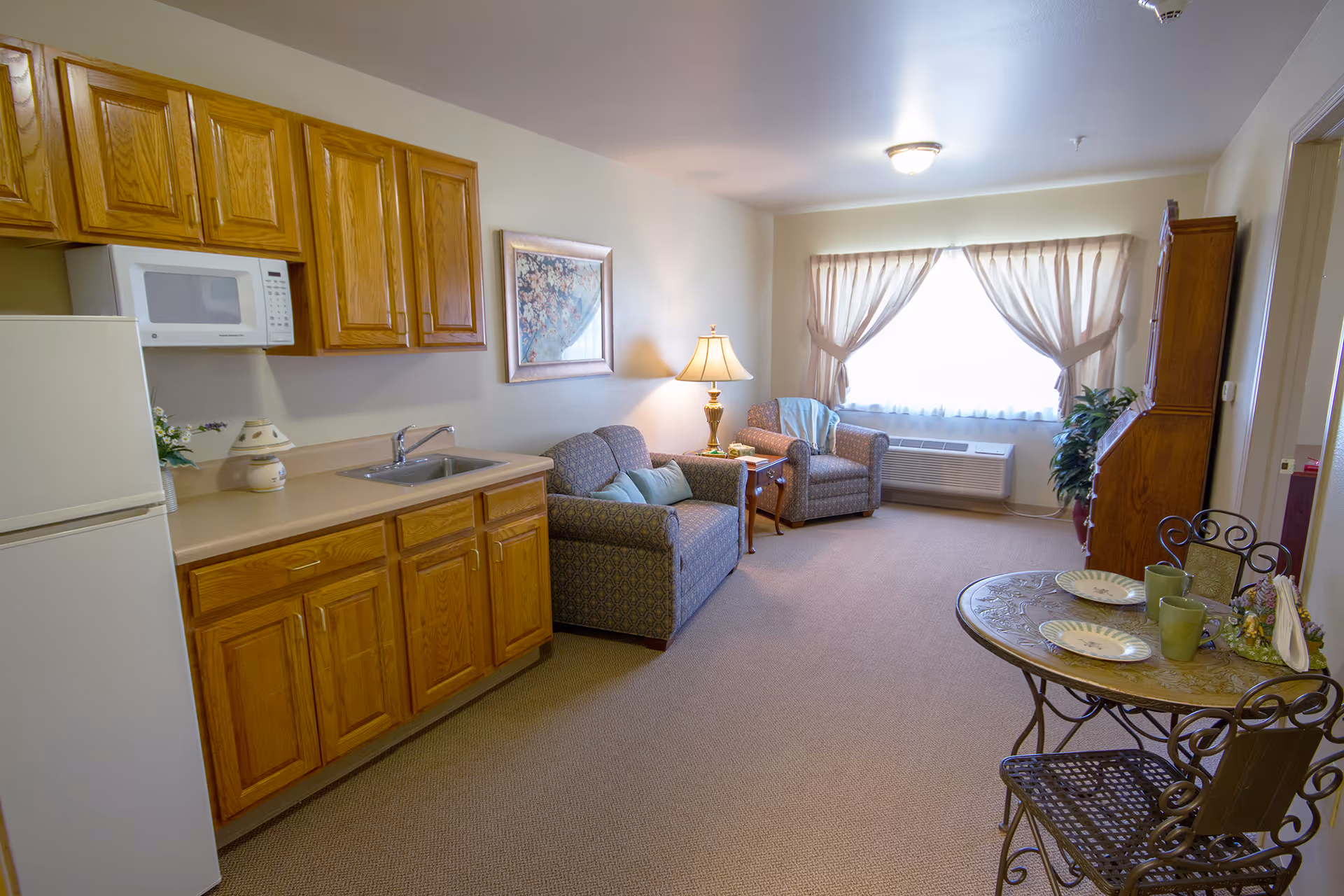 A cozy living area in a senior living facility featuring a small kitchenette with wooden cabinets, a white refrigerator, and a microwave. The room includes a patterned loveseat and armchair with a side table and lamp between them. A round dining table with two chairs is set with plates and cups. A large window with sheer curtains allows natural light to fill the room.