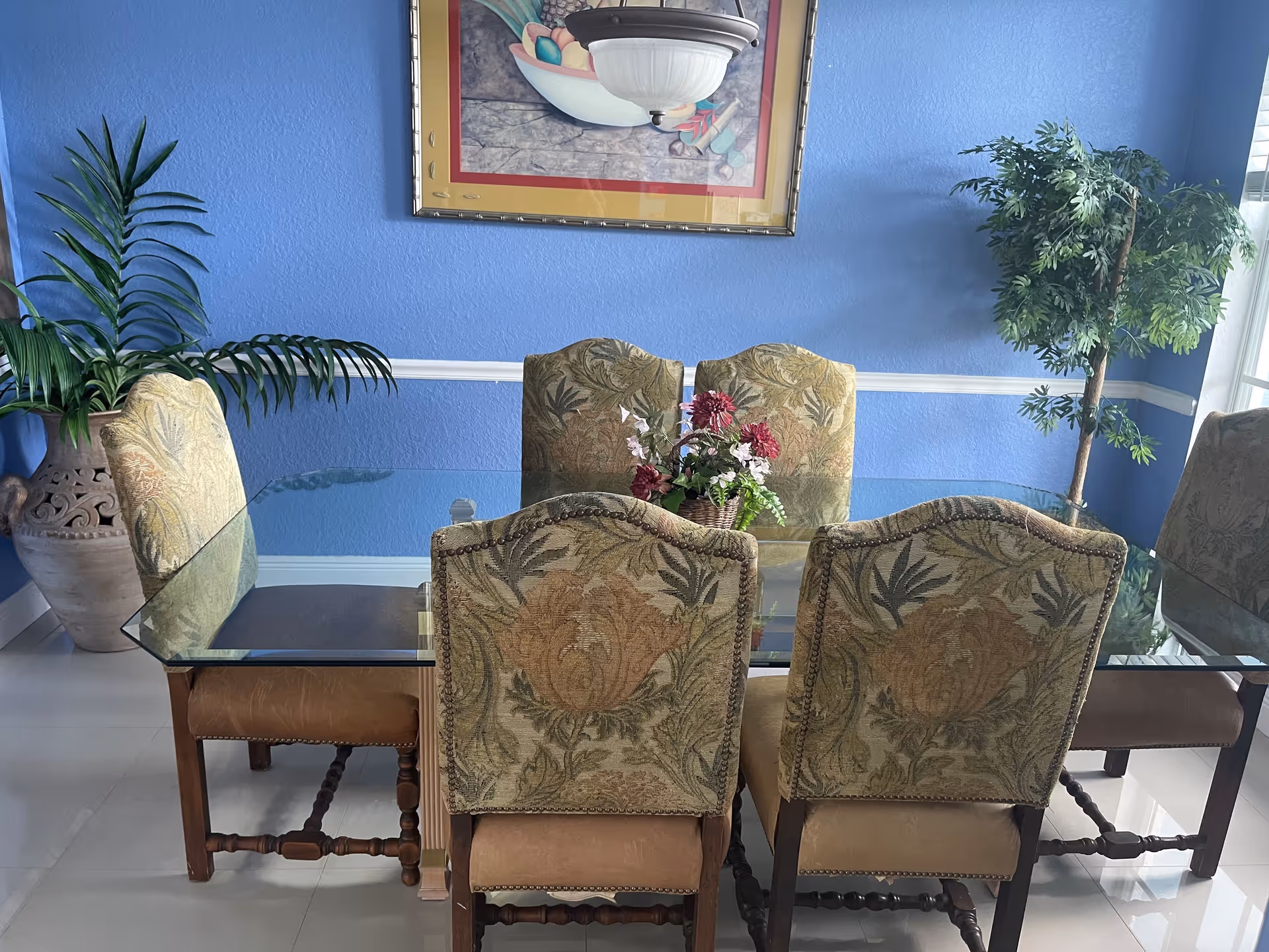 A dining room with a glass-top table surrounded by six upholstered chairs with floral patterns. The walls are painted blue with white trim, and there are two large potted plants on either side of the table. A framed painting and a ceiling light fixture are visible above the table.