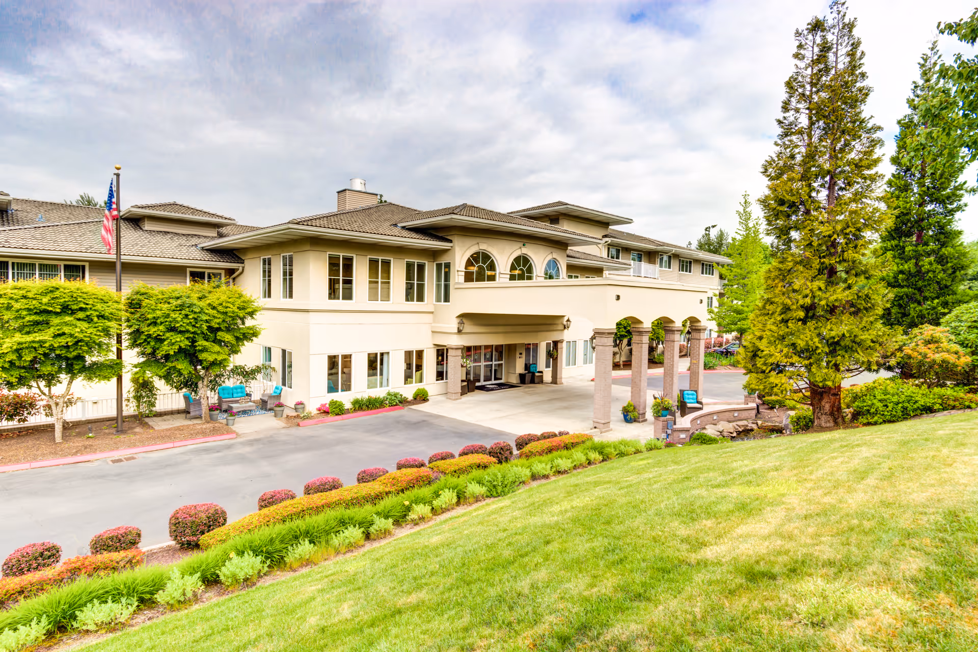 Exterior view of a senior living facility building with a covered entrance supported by columns, surrounded by well-maintained landscaping including green grass, bushes, and trees under a cloudy sky.