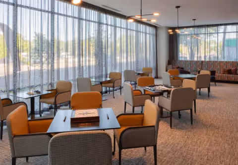 A spacious, well-lit common area with multiple tables and chairs arranged for games and socializing. Large windows with sheer curtains allow natural light to fill the room. Some tables have board games on them, and there is a patterned cushioned bench along the far wall.