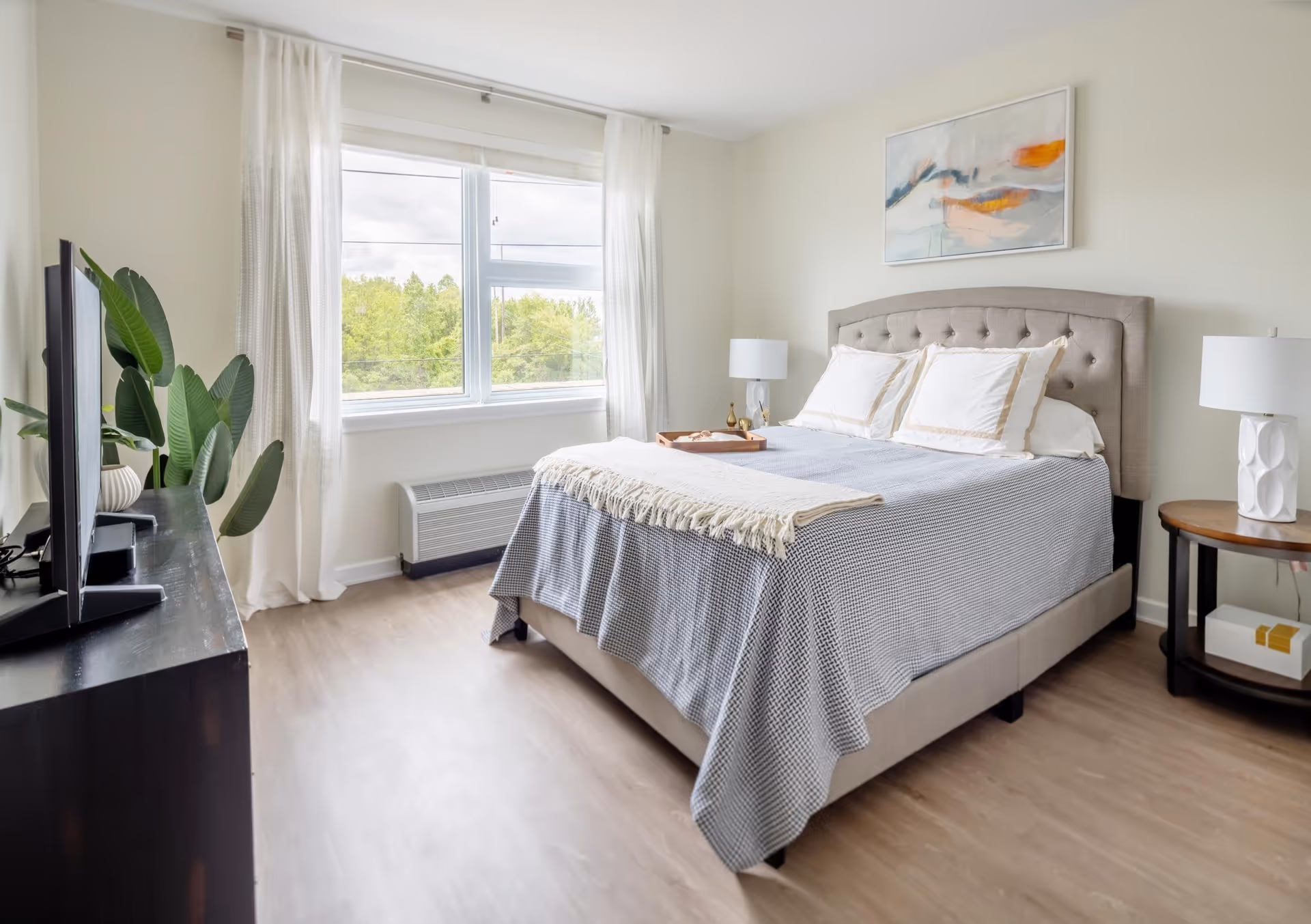 A bright bedroom with a large window covered by sheer white curtains, a bed with a tufted beige headboard, white pillows, and a gray blanket. There is a wooden nightstand with a white lamp on the right side of the bed, a dark wooden dresser with a flat-screen TV on the left, and a large green plant next to the dresser. A colorful abstract painting hangs on the wall above the bed.