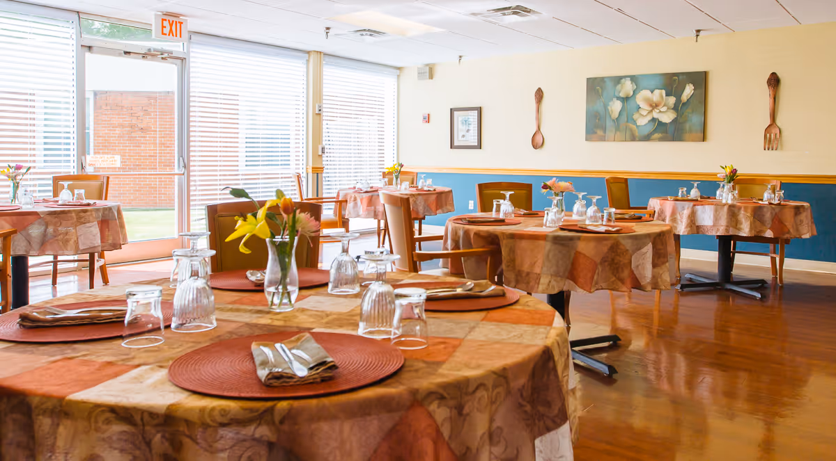 A bright dining room with round tables covered in patterned tablecloths, each set with placemats, napkins, utensils, and upside-down drinking glasses. Small flower vases decorate the tables. The room has large windows with blinds allowing natural light to enter, wooden chairs, and a painting of flowers on the wall along with decorative wooden utensils.