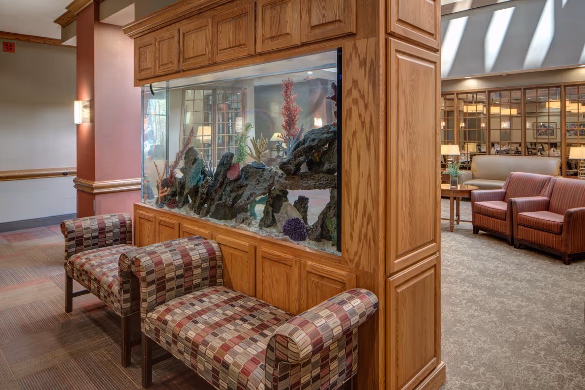 Interior view of a senior living facility lounge area featuring a large built-in aquarium with coral decorations. In front of the aquarium are two patterned upholstered benches. In the background, there are additional seating areas with armchairs and a sofa, along with wooden framed glass partitions and soft lighting.