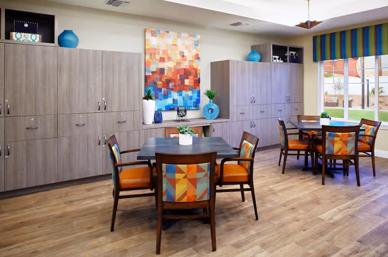 A bright and modern common area with two square tables surrounded by colorful cushioned chairs. The room features wooden flooring, large cabinets along the wall, decorative vases, a colorful abstract painting, and a large window with a striped valance overlooking an outdoor space.
