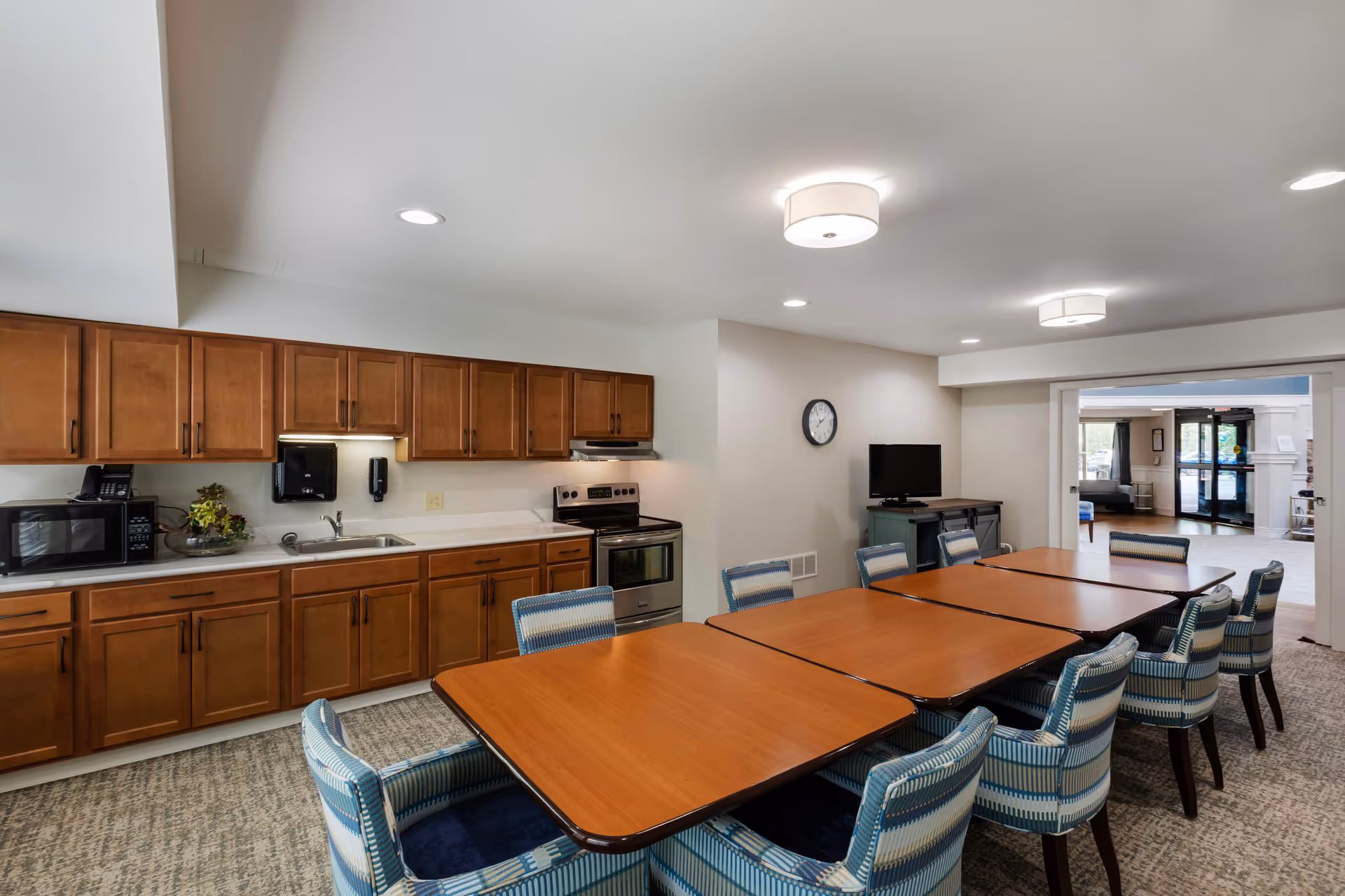 A communal kitchen and dining area in a senior living facility featuring wooden cabinets, a microwave, a stove, a sink, and multiple tables with blue patterned chairs. The room is well-lit with ceiling lights, and a small TV is mounted on a cabinet against the wall. An open doorway leads to another room with large windows and seating.