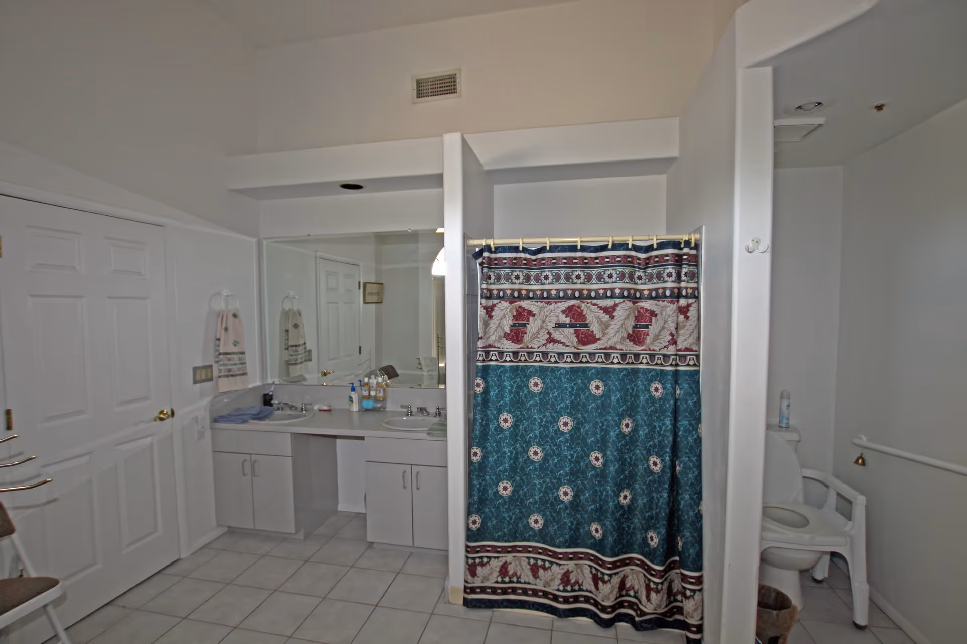 Spacious bathroom with double-sink vanity and large mirror, tiled floor, shower with patterned curtain, and a toilet with a commode chair.