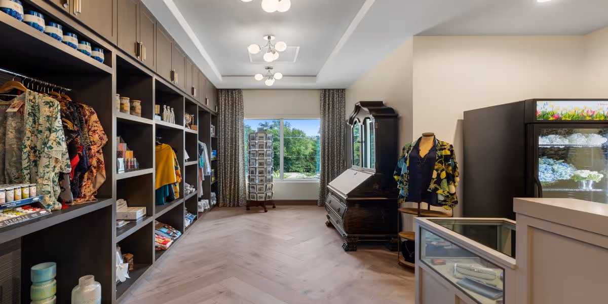 Interior view of a retail shop area within a senior living facility, featuring shelves stocked with clothing, canned goods, and other items on the left. On the right, there is a glass display case, a mannequin dressed in a floral shirt, and a refrigerated display with flowers. A large window with curtains at the end of the room lets in natural light and shows greenery outside.