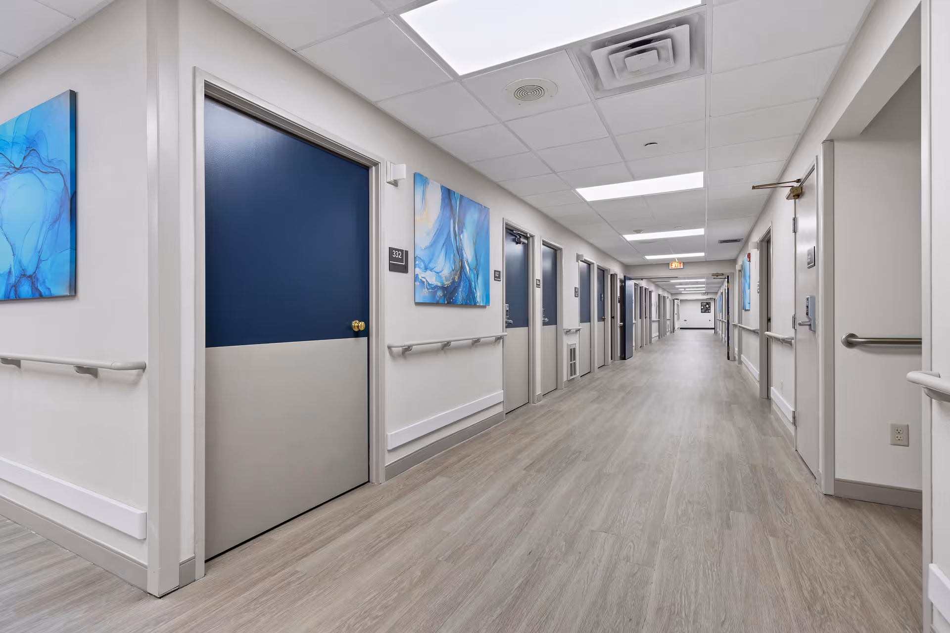 Long, well-lit interior hallway in a senior living facility with numbered blue doors, handrails, and artwork on the walls.