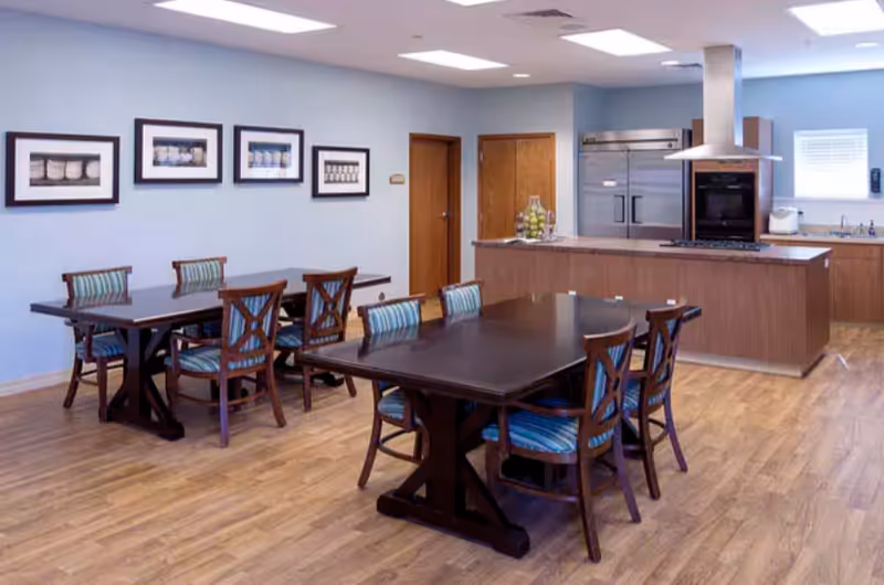 Dining area with dark wooden tables and striped upholstered chairs facing a modern kitchen island and stainless appliances.