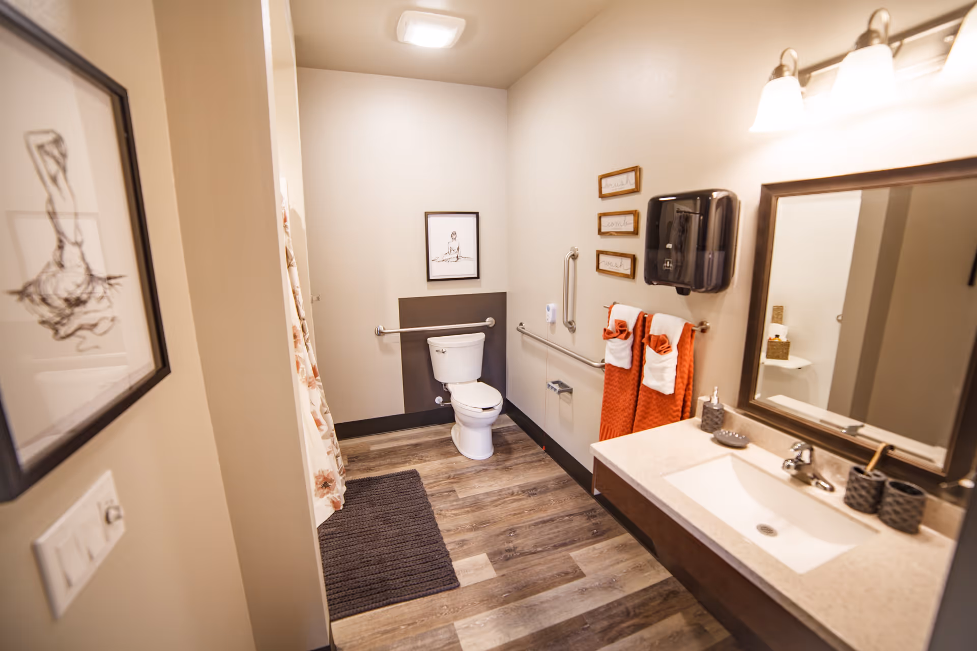 A clean and well-lit bathroom with a toilet, a sink with a large mirror above it, and a shower curtain partially visible. The bathroom has wooden flooring, a dark gray bath mat, and orange towels hanging on a towel rack. There are safety grab bars near the toilet and a paper towel dispenser on the wall. Two framed artworks are visible on the walls.