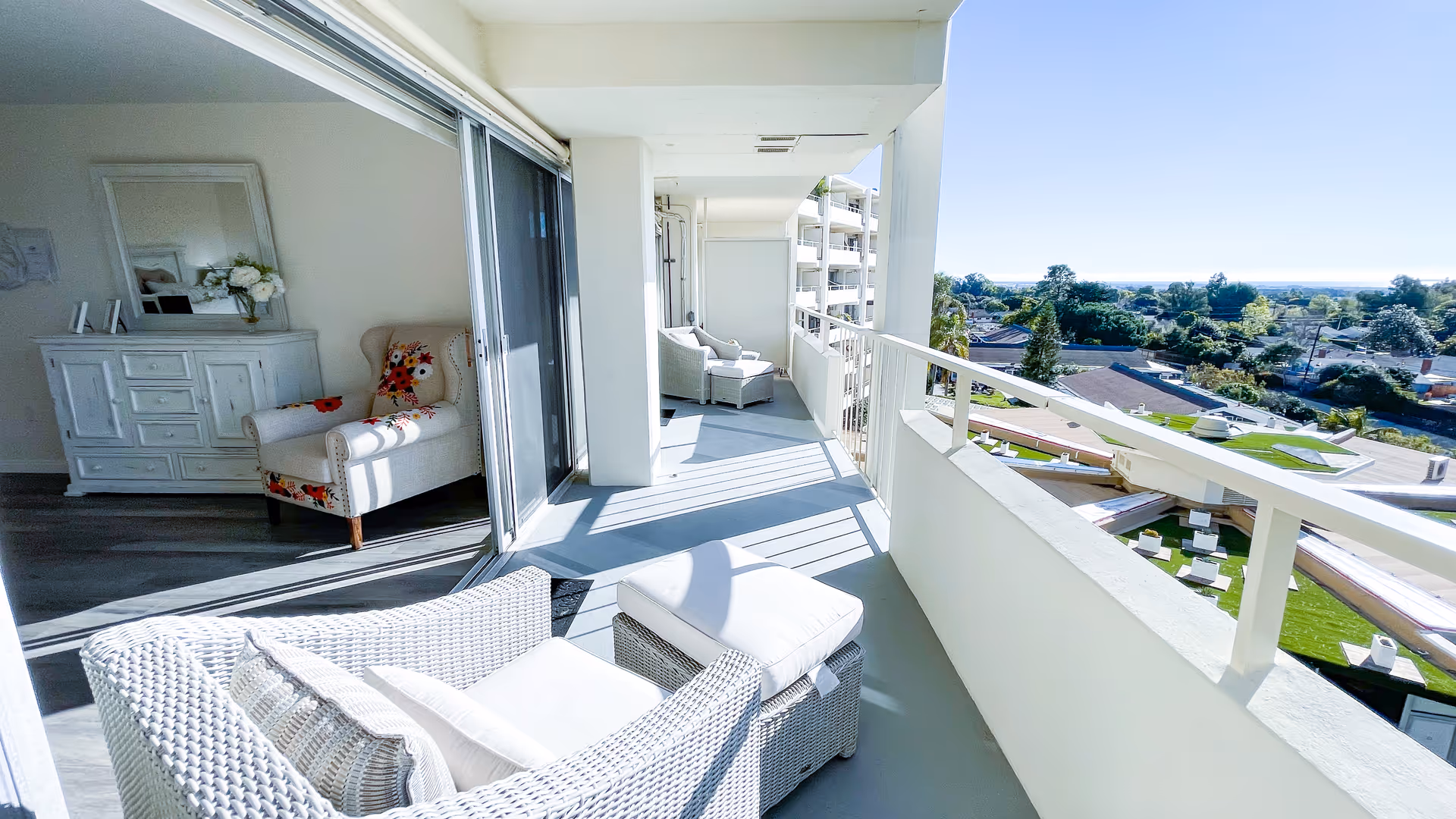 A bright balcony with white wicker chairs and cushions, overlooking a residential neighborhood with trees and houses. Inside, through sliding glass doors, a room with a white dresser, a mirror, and a floral-patterned armchair is visible.