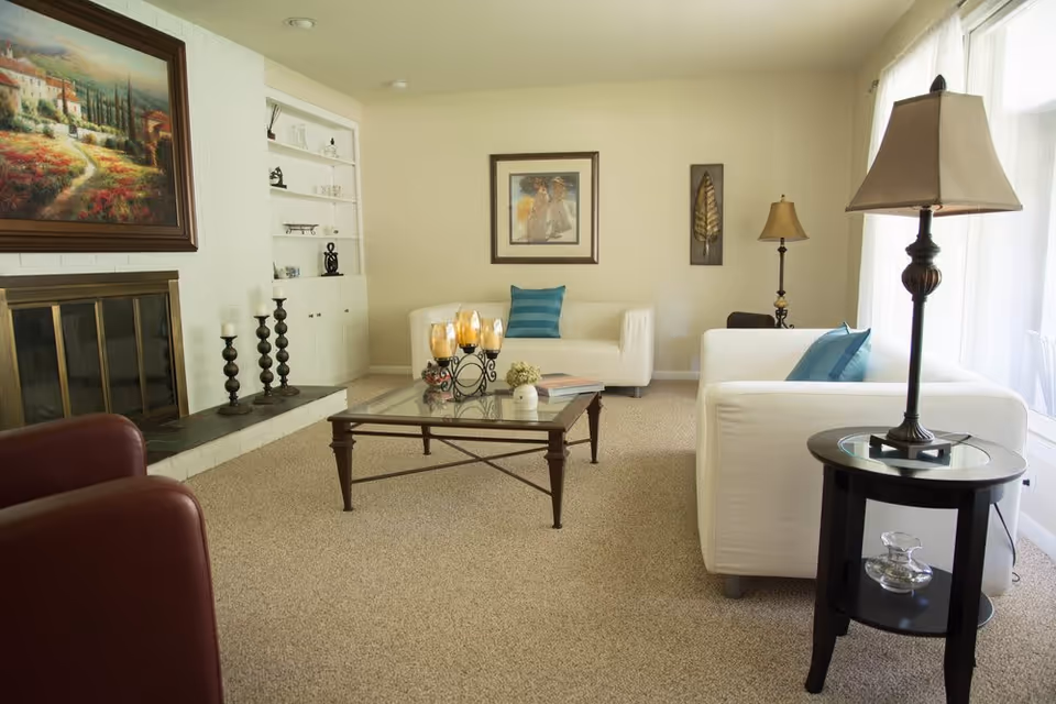 A bright living room with beige carpet, two white sofas with blue pillows, a glass coffee table with decorative items, a fireplace with candle holders, built-in shelves, and framed artwork on the walls. There are two table lamps near the sofas and large windows with sheer curtains letting in natural light.