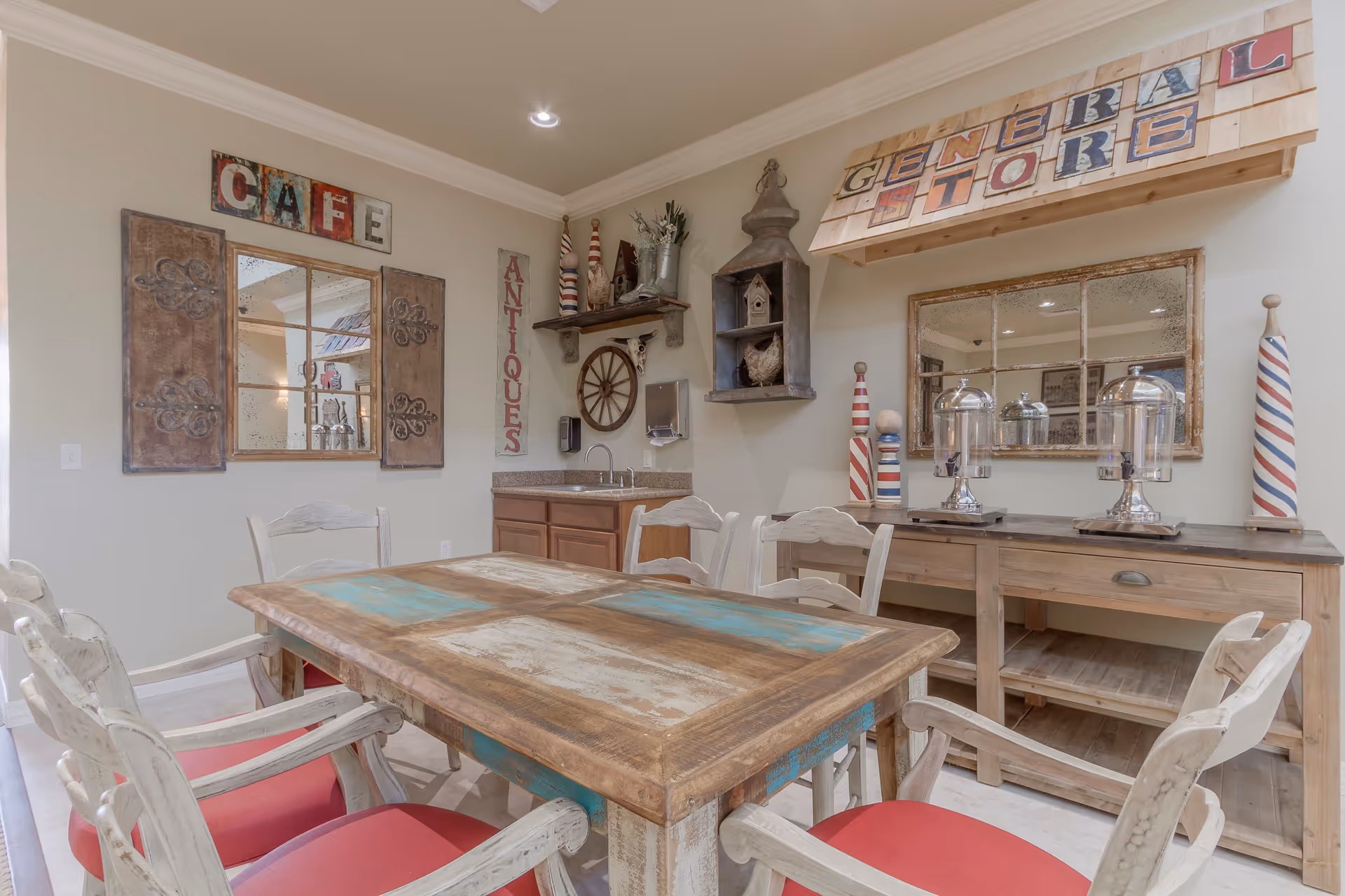 A rustic dining area with a wooden table and six chairs with red cushions. The walls are decorated with vintage-style signs reading 'CAFE', 'ANTIQUES', and 'GENERAL STORE'. There are decorative items including a wooden wheel, a birdhouse, and striped barber pole-style ornaments. A wooden sideboard with glass dome covers and a sink with a countertop are also visible.