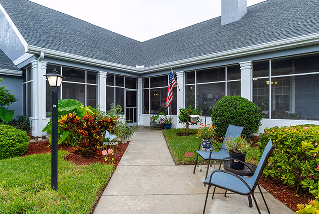 Pathway leading to a screened porch entrance with an American flag, landscaped beds and outdoor chairs.