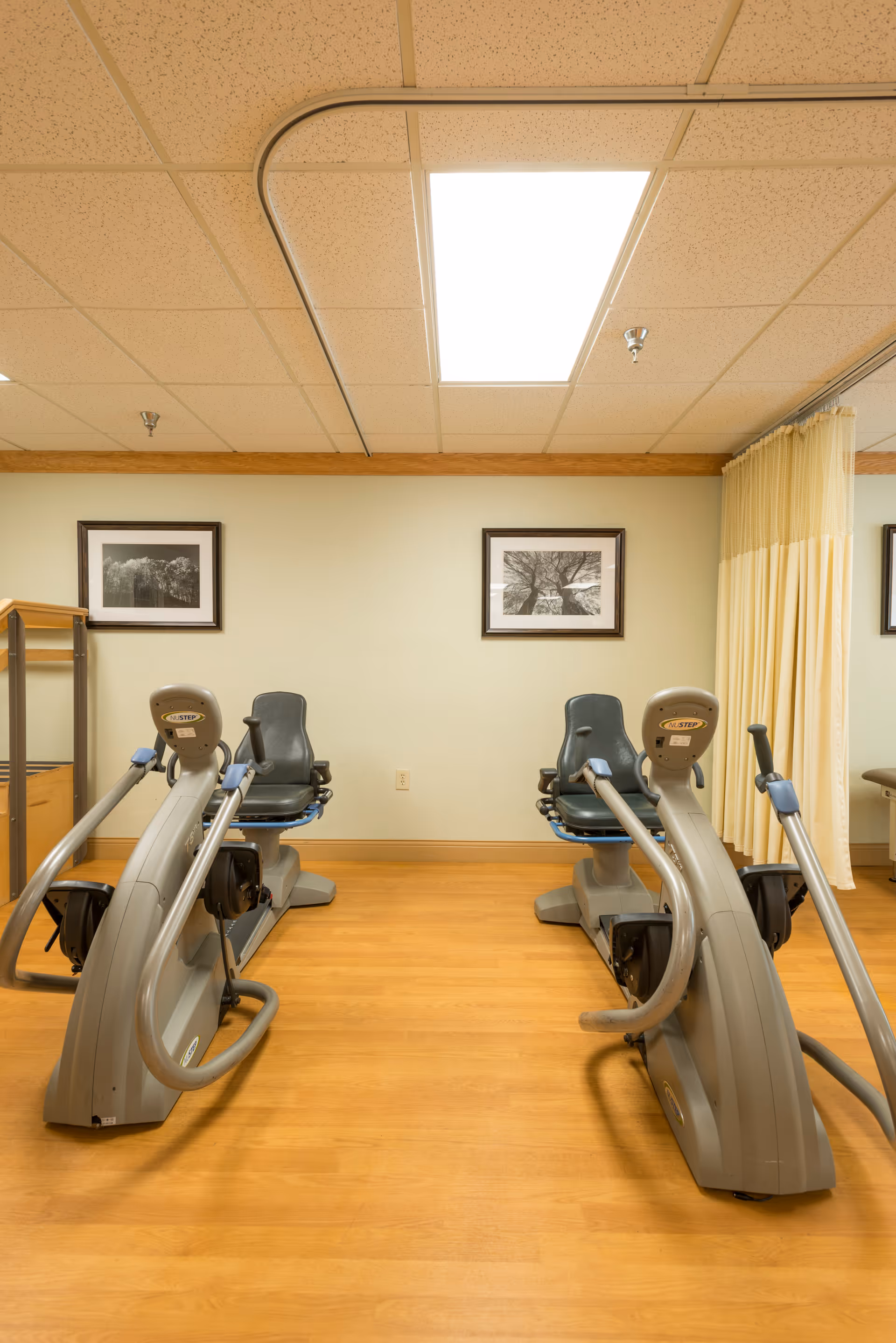 A small exercise room with two recumbent stationary bikes facing a wall with two framed black and white photographs. The room has a wood floor, beige walls, a drop ceiling with fluorescent lighting, and a yellow privacy curtain on the right side.