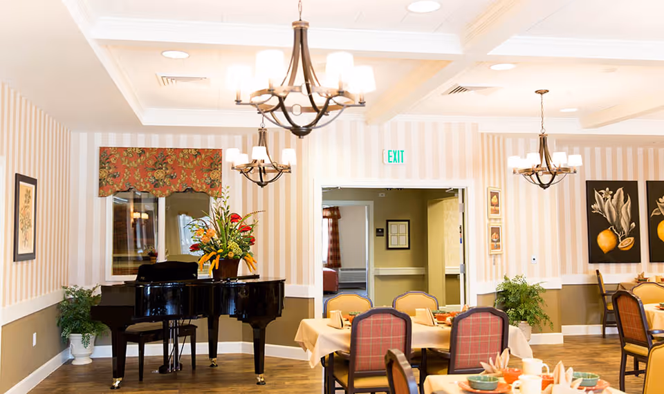 A bright dining room with tables set for a meal, featuring a grand piano with a floral arrangement on top, striped wallpaper, framed artwork, and elegant chandeliers hanging from the ceiling.