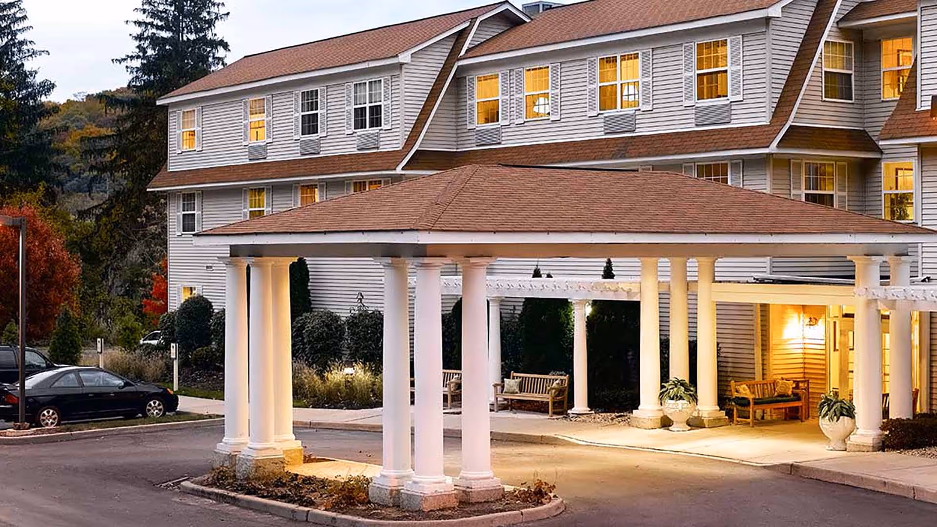 Exterior view of Atria Briarcliff Manor senior living facility at dusk, showing a covered entrance with white columns, benches, potted plants, and a multi-story building with lit windows in the background.