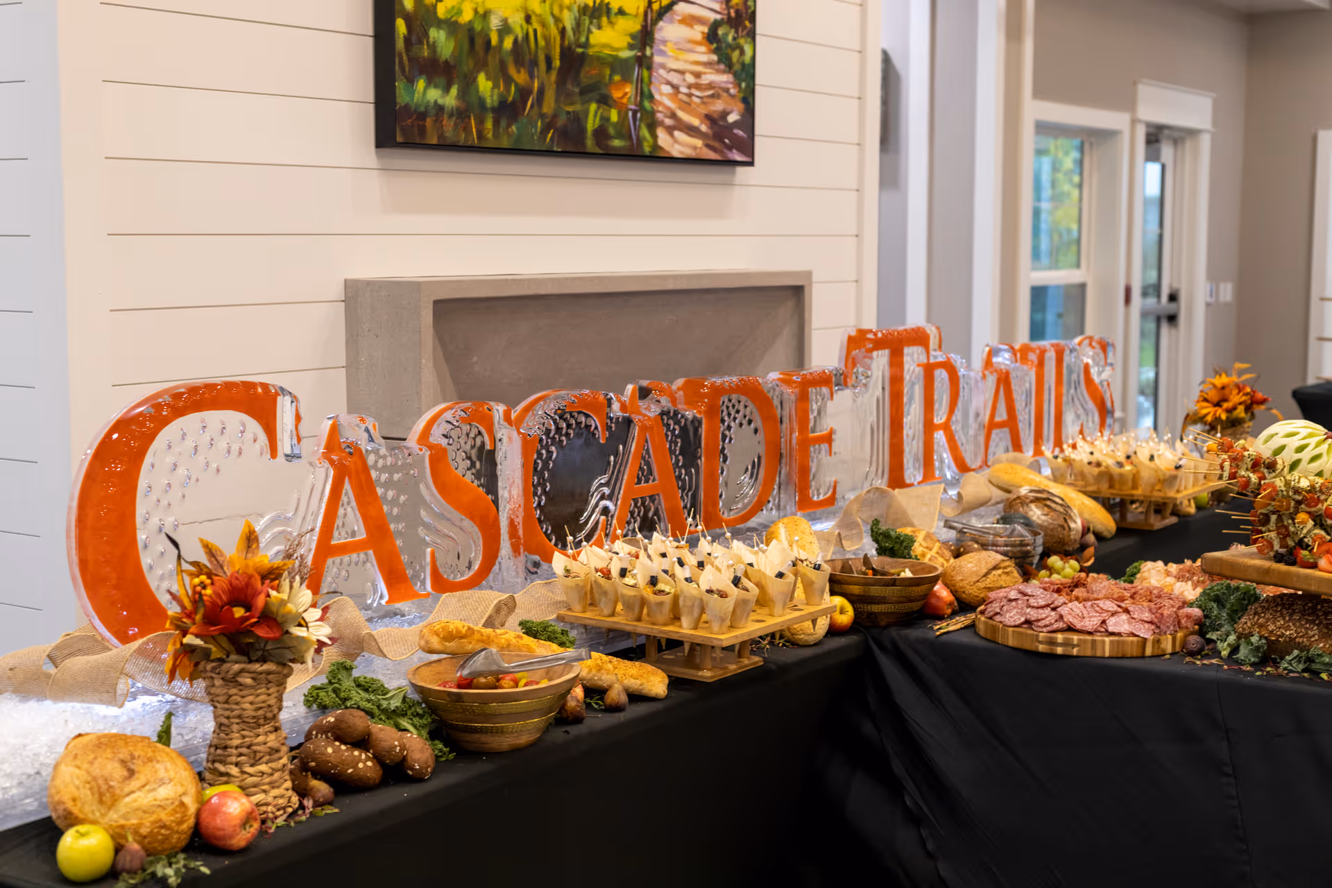 A buffet table set up with various breads, meats, fruits, and appetizers in a well-lit indoor space. Behind the table is a large ice sculpture spelling out 'CASCADE TRAILS' in orange and clear ice. The room has light-colored walls, a painting, and windows letting in natural light.