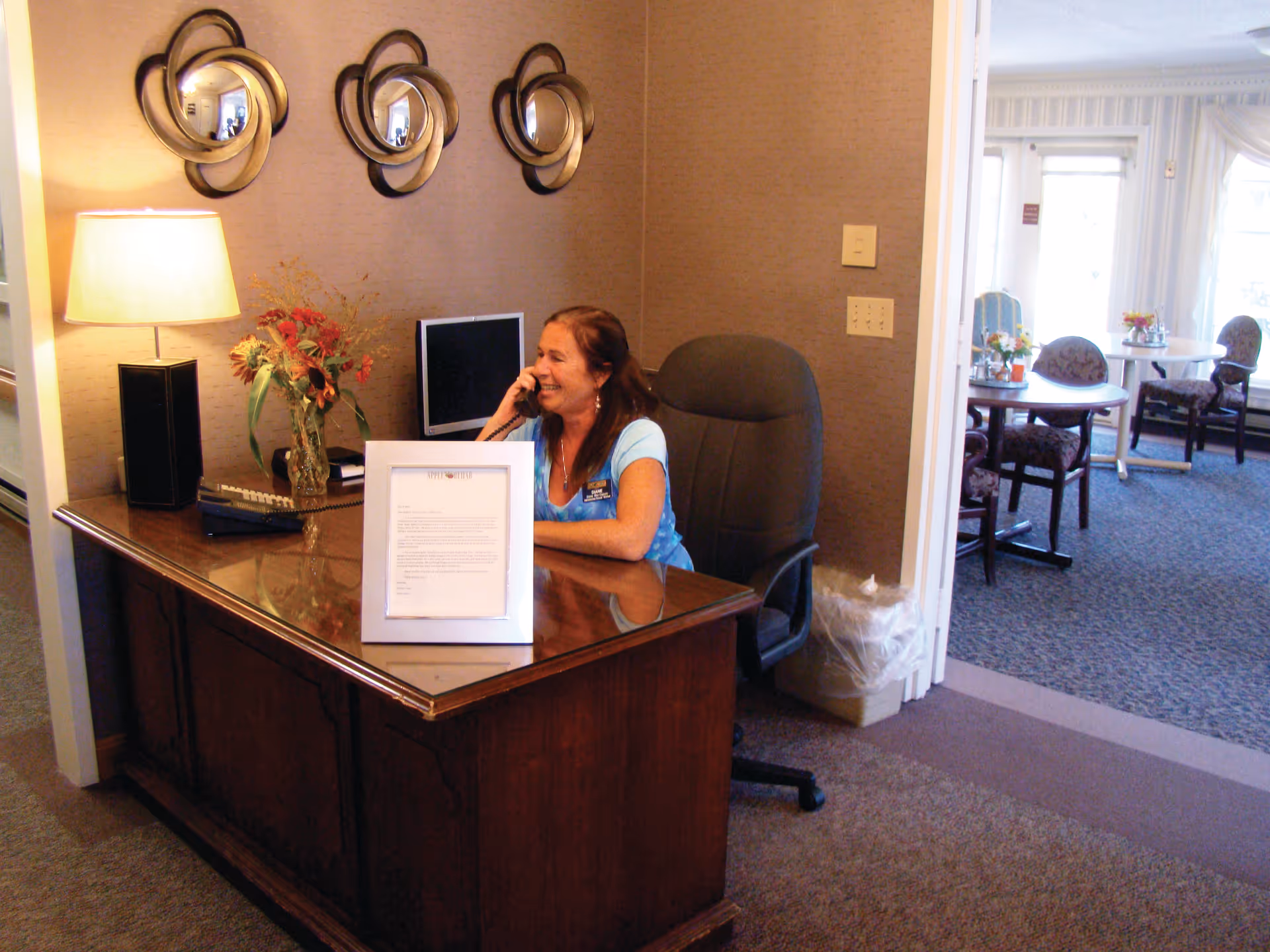 A woman sitting at a wooden reception desk in an office area, talking on the phone. The desk has a lamp, a vase with flowers, a computer monitor, and a framed document. Behind her is a beige wall with three decorative circular mirrors. In the background, there is a room with tables and chairs near large windows with curtains.