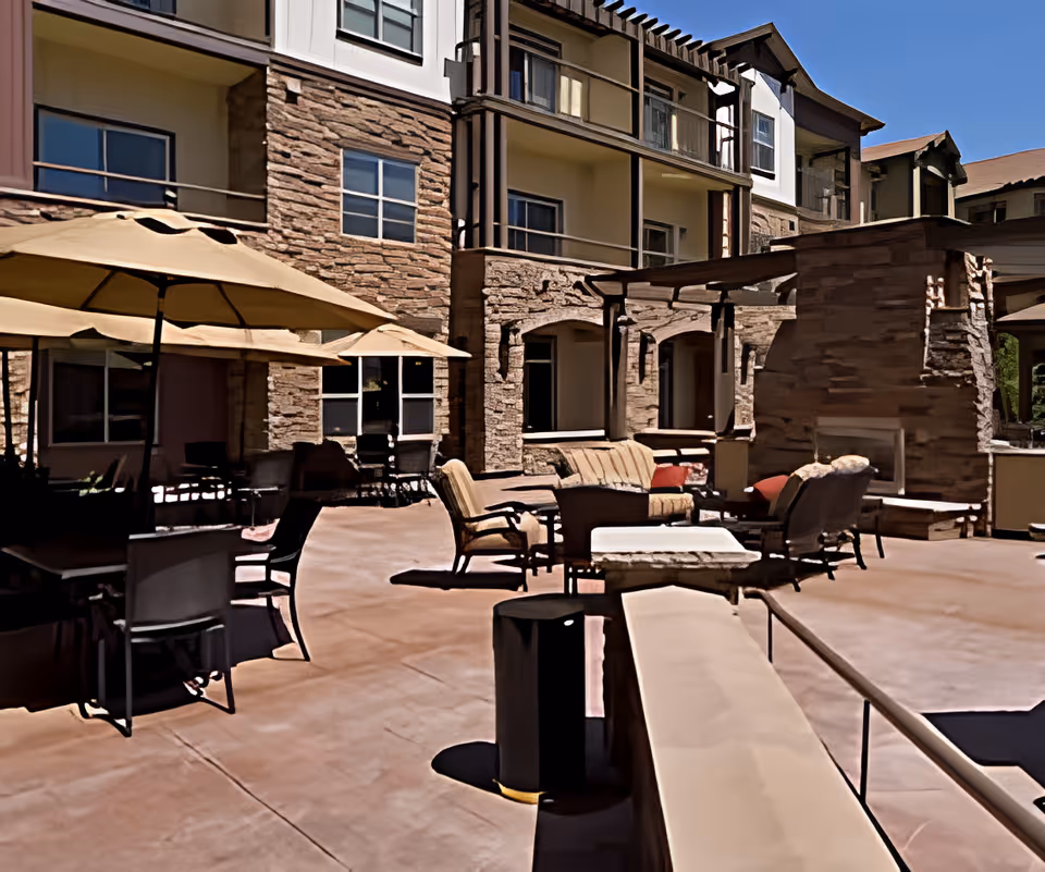 Outdoor patio area of a senior living facility with cushioned chairs, sofas, tables, large umbrellas for shade, and a stone fireplace. The building exterior features stone and stucco walls with multiple windows and balconies under a clear blue sky.