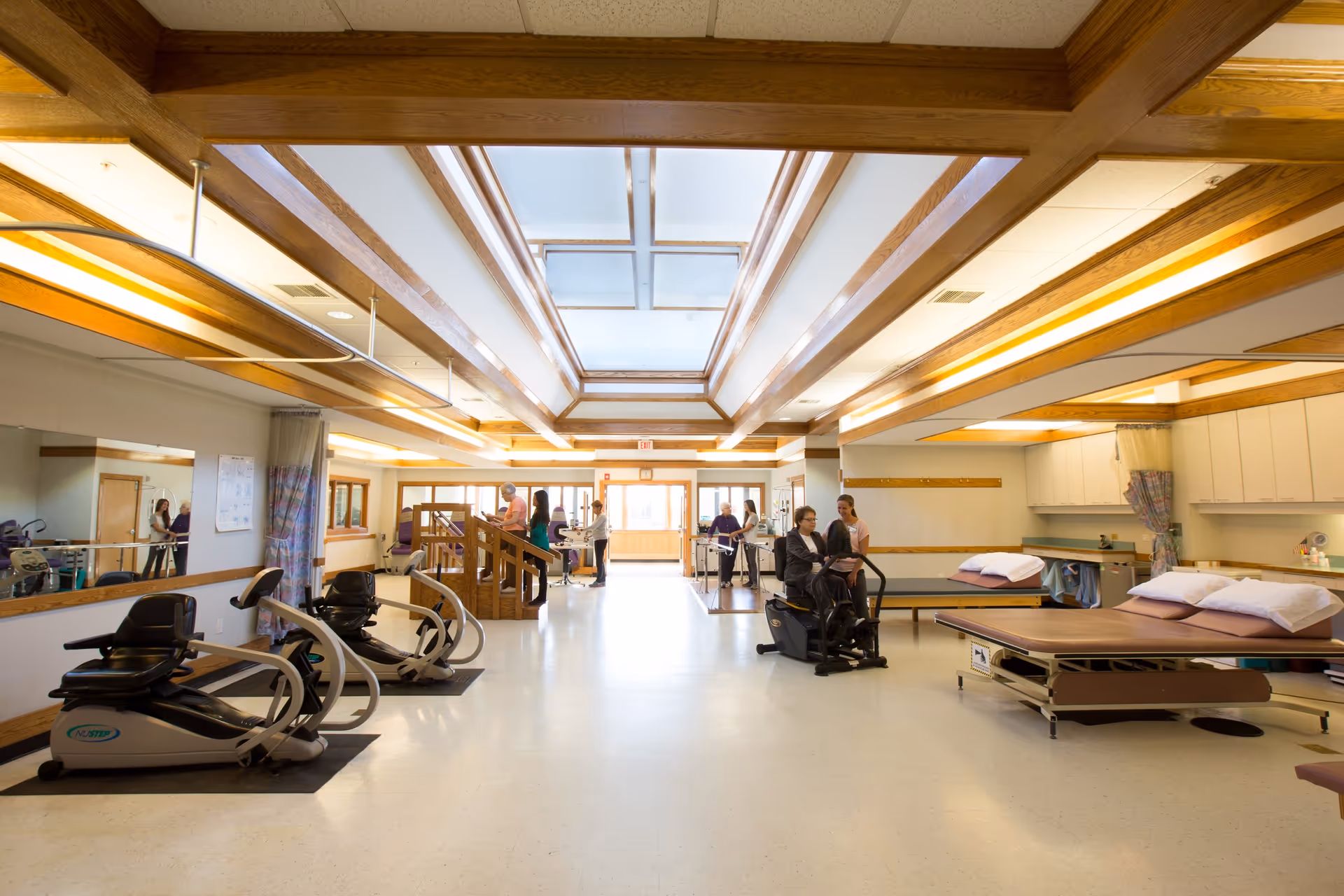 A spacious rehabilitation room with exercise equipment, therapy tables, and parallel bars. Several people are engaged in physical therapy activities, assisted by staff. The room features wooden ceiling beams and a large skylight providing natural light.