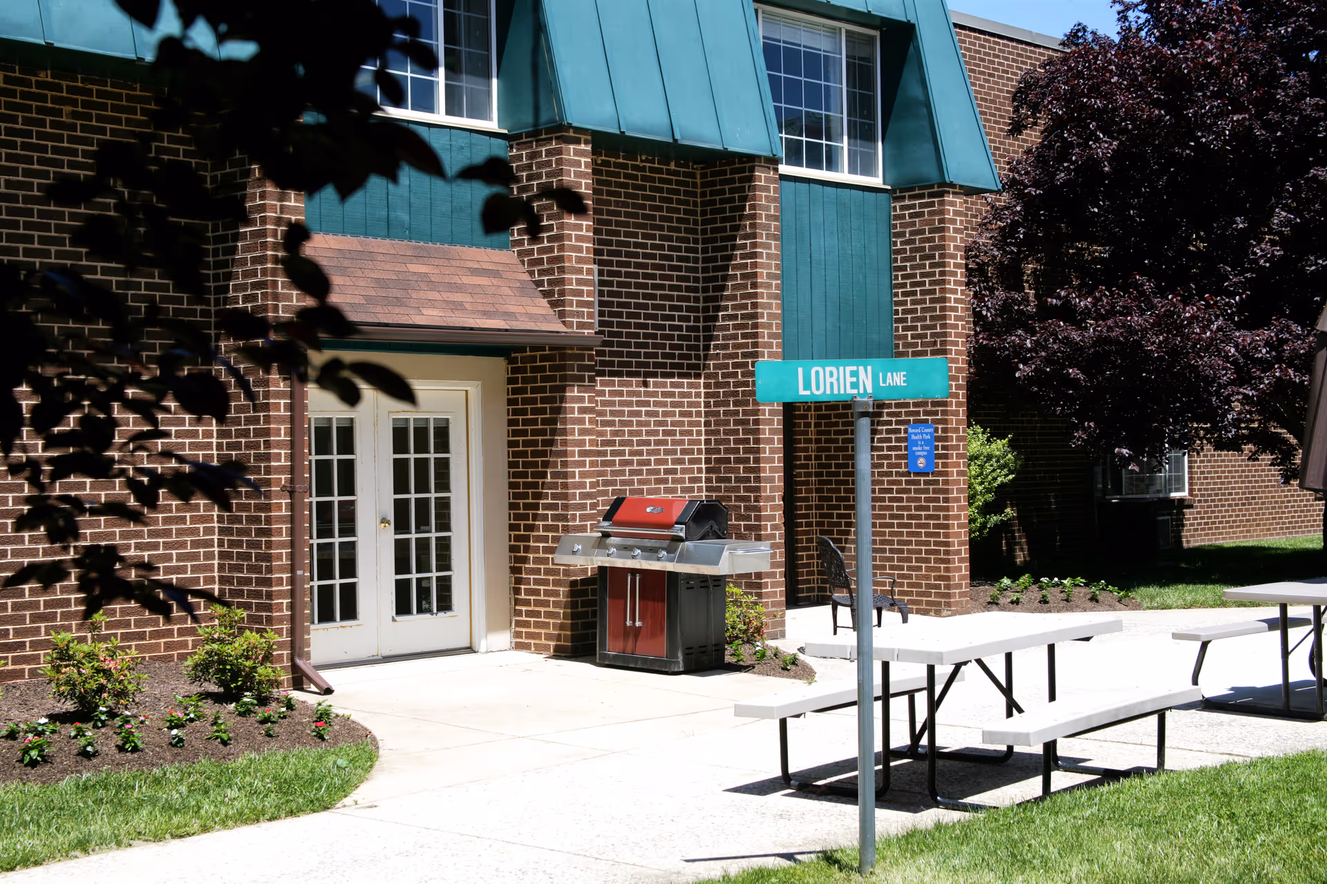 Outdoor courtyard at a brick senior living building with picnic tables, a grill, and a 'Lorien Lane' sign.