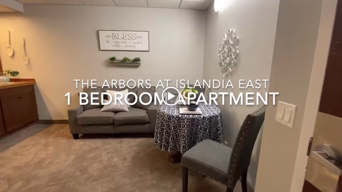 Interior view of a one-bedroom apartment at The Arbors Assisted Living Communities at Islandia East, showing a small living area with a gray loveseat, a round table covered with a patterned tablecloth, and a gray upholstered chair. The walls are decorated with a framed sign that says 'BLESS' and a metal wall art piece. Part of a kitchen counter is visible on the left side.