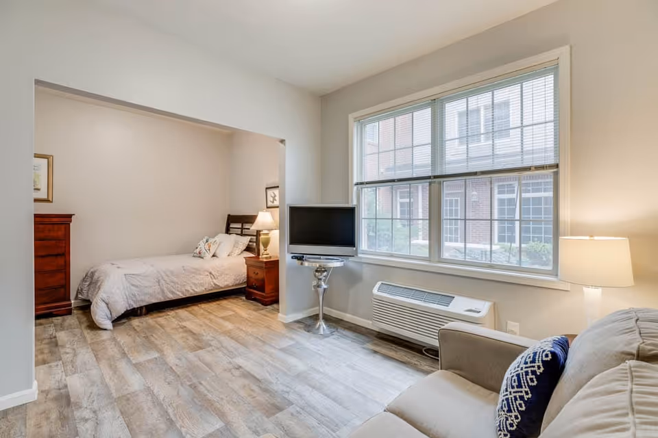 Studio-style assisted living interior with a sofa and TV by a large window and a bed in an alcove.